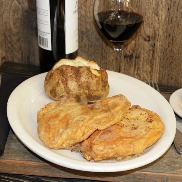 Two golden-brown pork chops and a baked potato on a white plate, with red wine in the background.