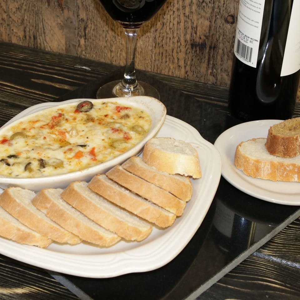 Baked dip with vegetables and cheese, served with sliced baguette and a glass of red wine.