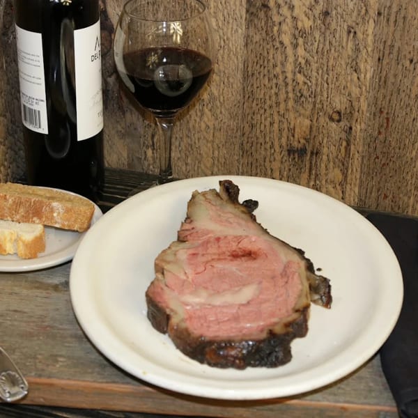A slice of prime rib, bread, and red wine on a rustic wooden table.