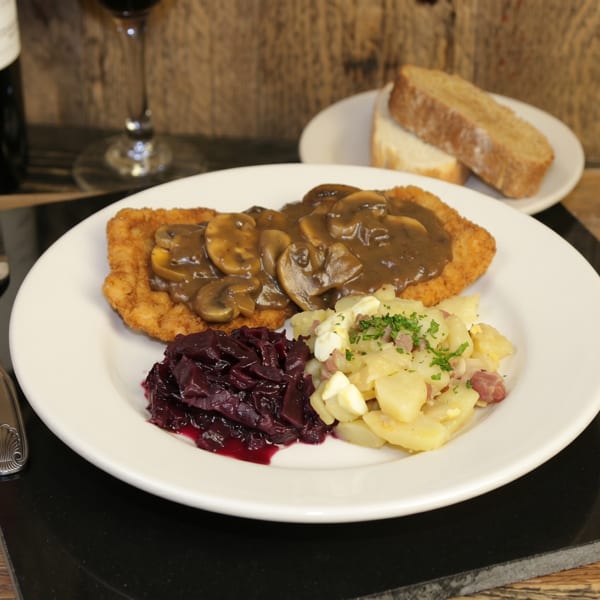 A plate of schnitzel with mushroom gravy, red cabbage, and potato salad, with wine and bread.